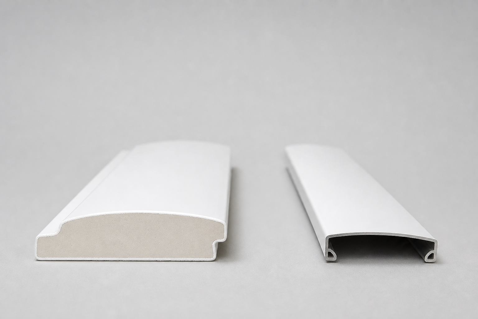 Side-by-side cross-section product photograph of two white plantation shutter louvres on a neutral grey backdrop, the left louvre is faux wood showing a solid uniform PVC core with a thick painted shell, the right louvre is aluminium showing a hollow extruded aluminium chamber with internal walls visible and a noticeably slimmer overall profile.