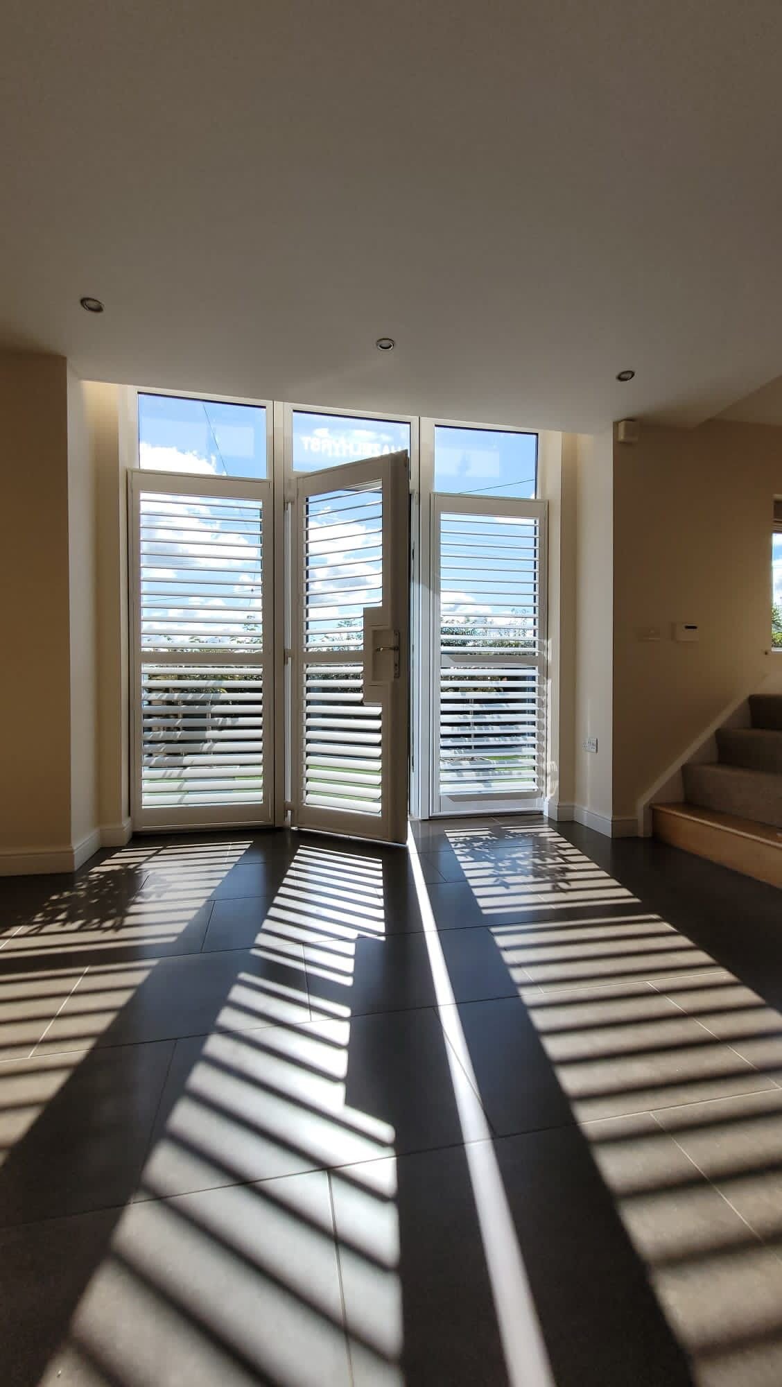 White plantation shutters on French doors with one leaf ajar, casting striped sunlight across a tiled floor.