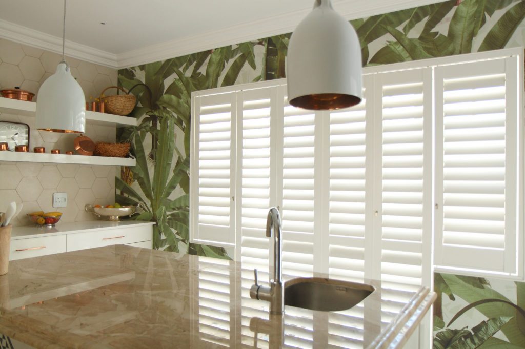 Four-panel white full-height plantation shutters on a wide modern kitchen window with louvres open for soft daylight
