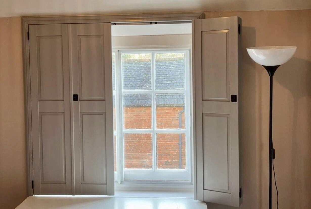 Platter-coloured solid panel shutters on a sash window, partially open, with black hinges and ring pulls