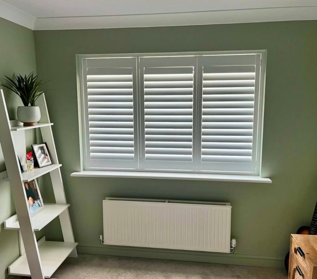 Close view of the Lowdham shutter install — three white panels with adjustable louvres in a sage-green bedroom