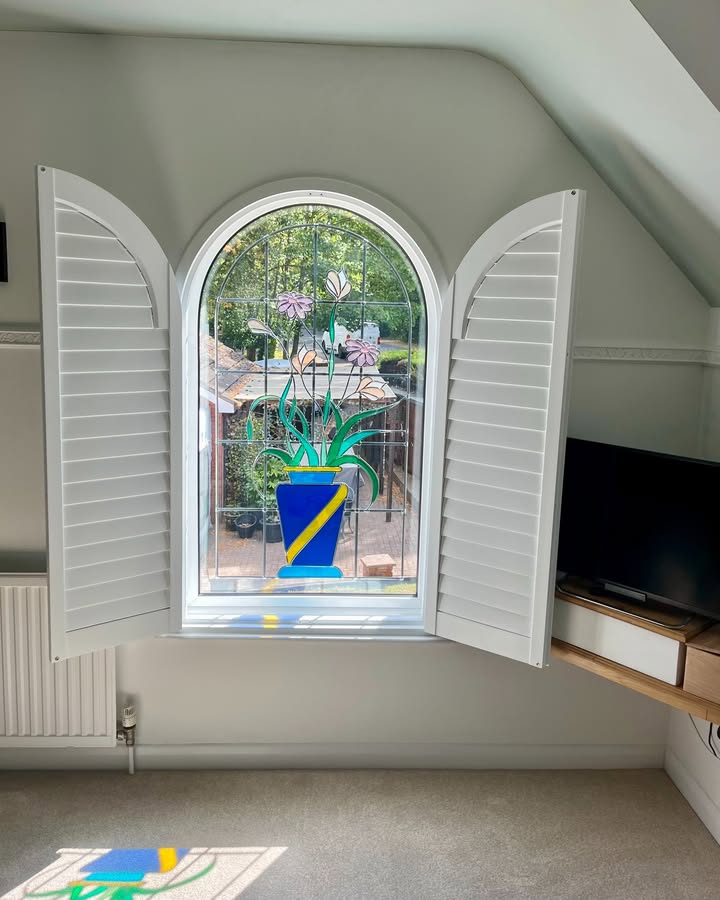 Arched white plantation shutters opened back to frame a stained-glass arched window in the Syston loft room